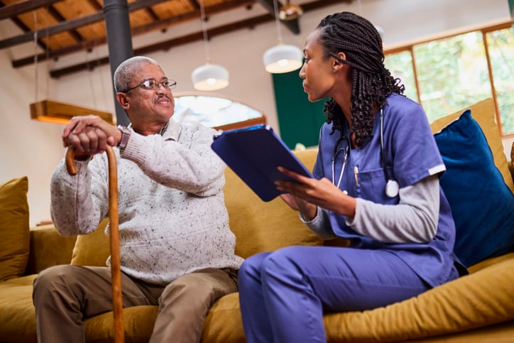A Black female nurse or caregiver in uniform sits with a senior African American man holding a cane, discussing his health and taking notes.